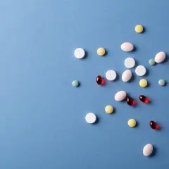 Różne tabletki i kapsułki, w tym białe, różowe, żółte i czerwone, rozsypane na niebieskim tle. Wśród nich widać kilka zielonych tabletek.