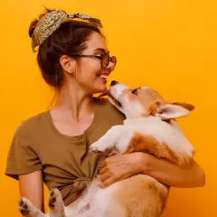 Uśmiechnięta dziewczyna z okularami i bandaną na głowie tuli corgi. Piesek liże ją po nosie. Wiele imion na N, jak Nela czy Nika, pasowałoby do tej słodkiej pary.